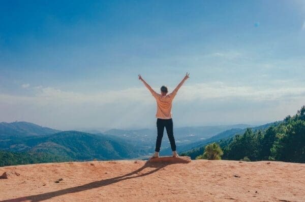 Man standing on the edge of mountain with hands in the air in front of blue sky and green mountain scenery