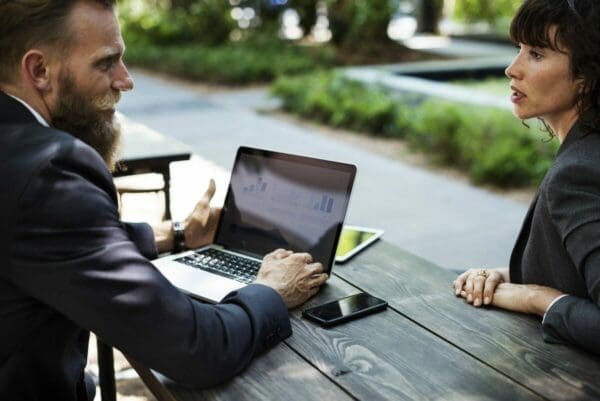 Two colleagues having a meeting outside on a bench with laptop, tablet and phone