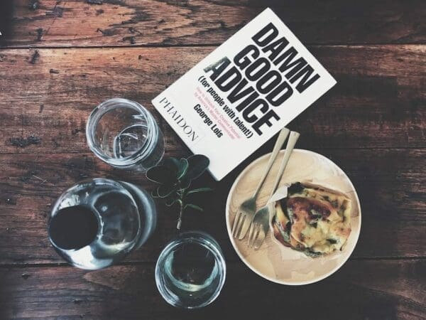 Book Damn Good Advice by George Lois on a wooden table with food, glass cups, and a bottle