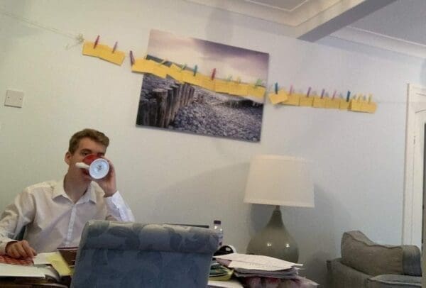 Young boy drinking coffee at his home desk used for revision with notes pinned on a line in the background