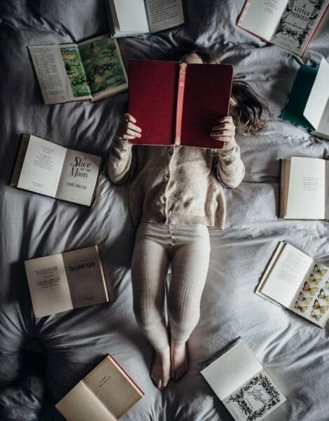 Little girl reading a red book while lying down on a soft bed with opened books surrounding her