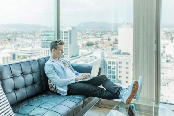Man using his laptop while sitting on a black sofa beside the windows of a tall building