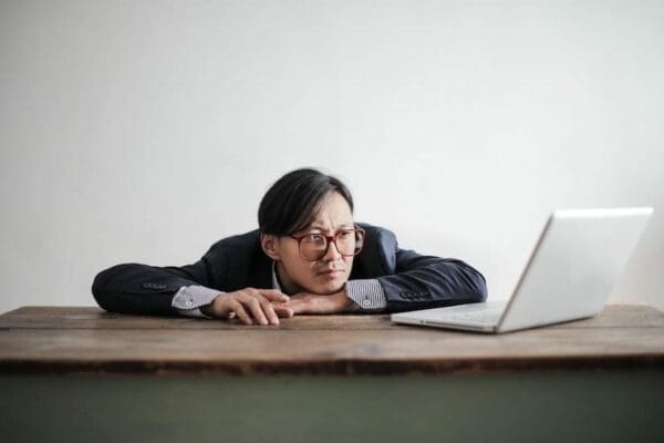 Man slumped on the table while staring at a boring virtual Zoom presentation on his laptop