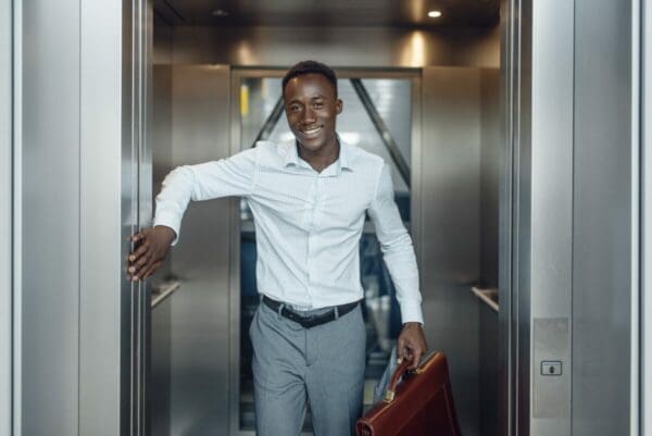 Young Businessman holding open the elevator door smiling about Buyer Tactics in Negotiation