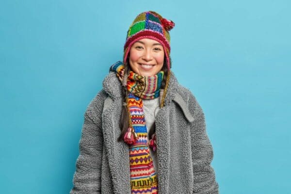 Young woman with a colourful hat and scarf on and a grey coat against a blue background