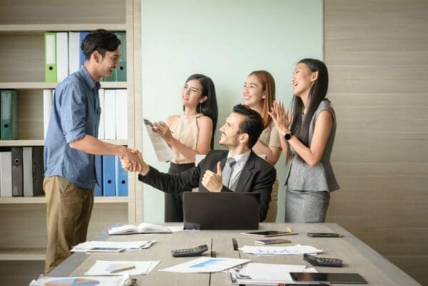 Manager congratulates male employee while female employees clap and cheer