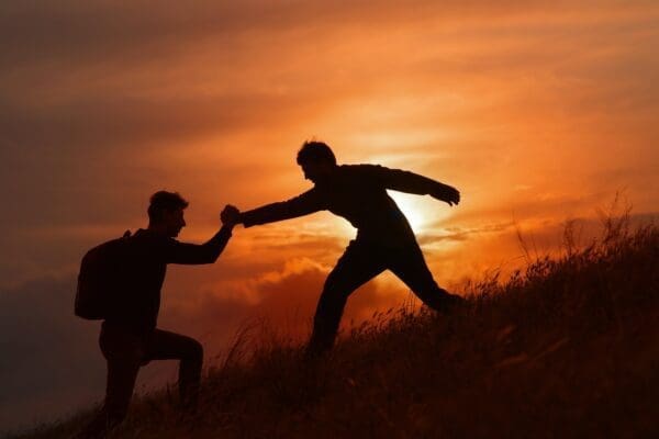 Silhouette of hiking teammate helping another teammate with sunset background