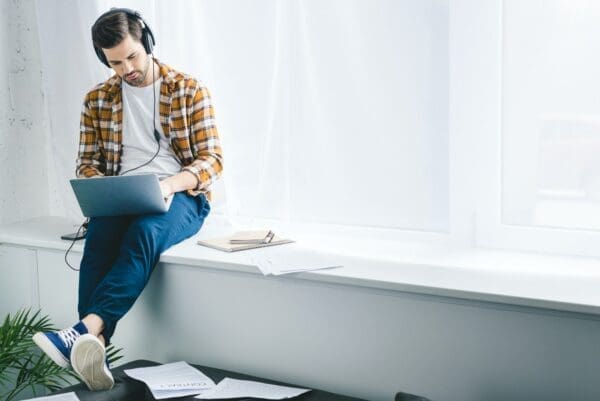 Remote worker using headphones and a laptop while sitting on a window sill