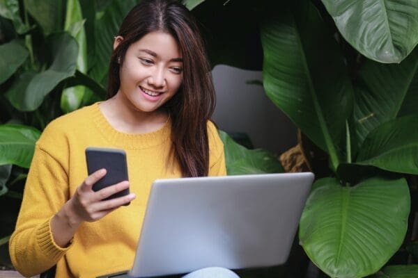 Woman on her laptop and phone with large green leaves in the background