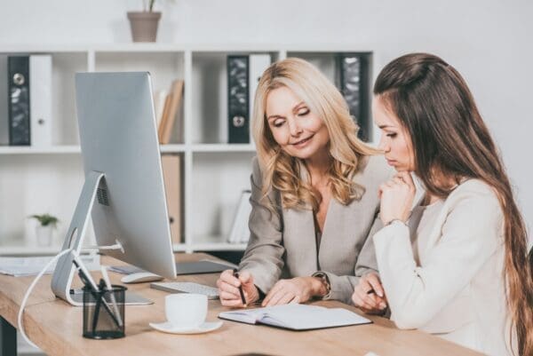 smiling female business mentor pointing at notebook and working with a young colleague in office