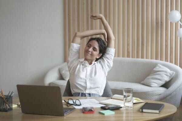Tired female remote employee stretching arms while sitting at desk with laptop for wellbeing