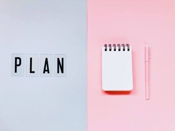 Plan Plan spelled on grey background beside a notepad on a pink background