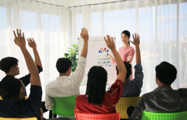 Businesswoman presenting from a whiteboard in a room of engaged employees raising their hands