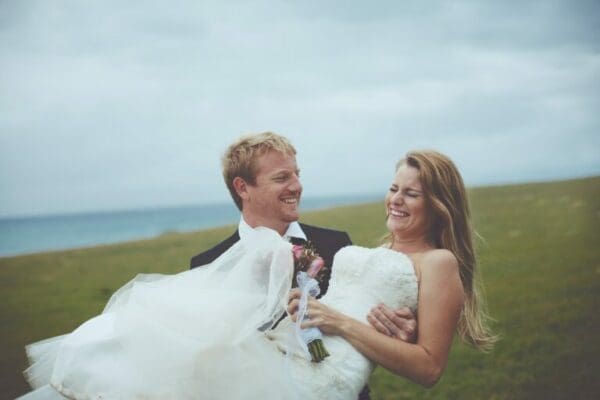 Happy Couple Just Married ISTP Groom carrying new bride across a field in his arms and both smiling