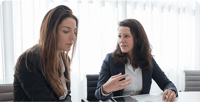 Two women engaged in a discussion at a table, with soft natural light filtering through sheer curtains in a modern office setting.