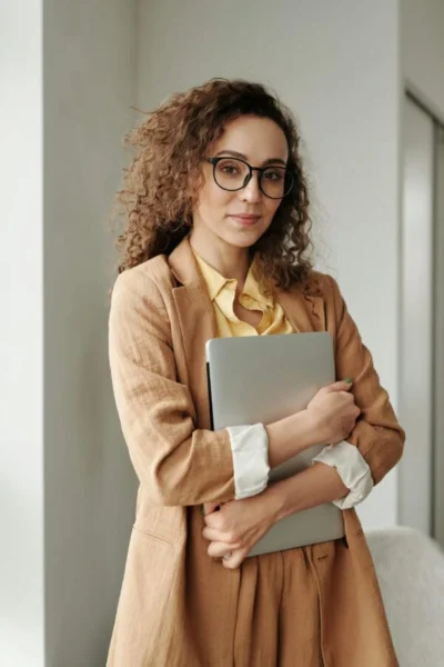 A woman in a tan blazer and cream blouse holds a laptop close, standing in a modern office space with soft, natural lighting.