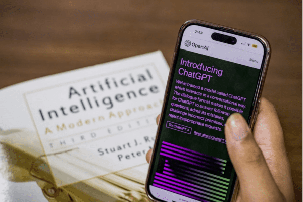A hand holding a phone with OpenAI's ChatGPT announcement page on screen, above an artificial intelligence book.