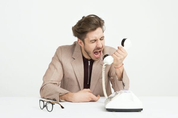 Man sitting at a white table with a white background shouting into a white telephone with a pair of glasses on the table